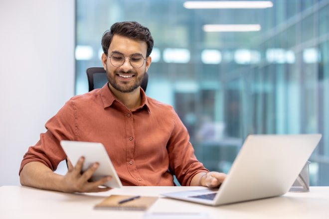 Confident young professional using a tablet and laptop in modern office environment for efficient