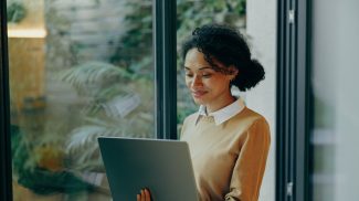 Female freelancer working laptop remotely while standing near window at home. Distance work concept
