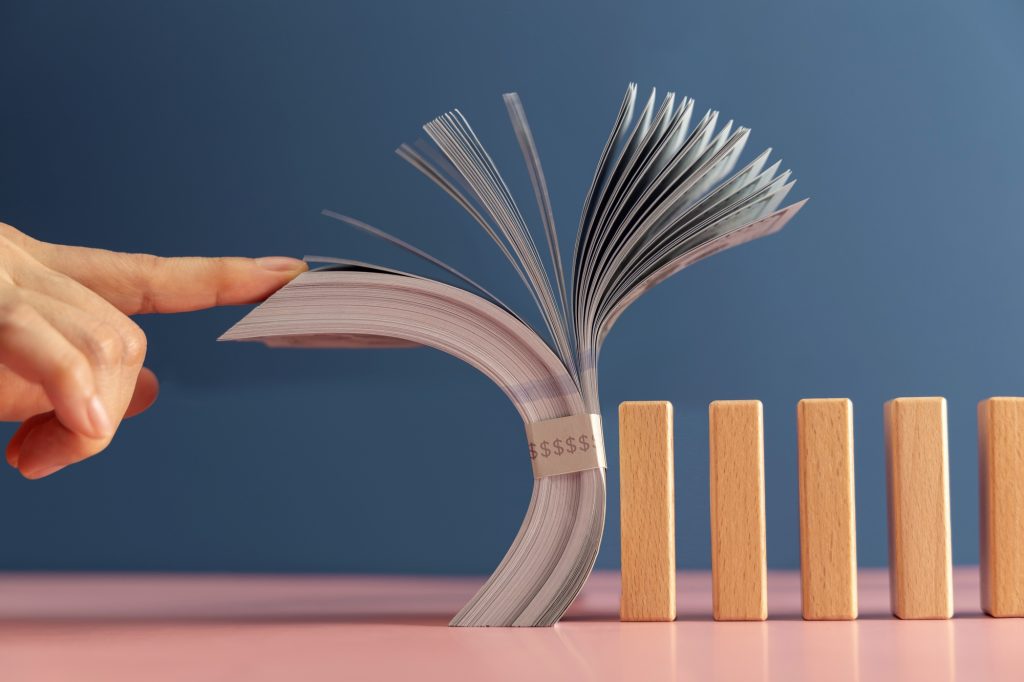 Finger Tapping US dollar stack onto dominos, symbolizing economic risk, global financial instability