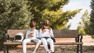 Two kids in astronaut and superhero costumes using mobile phone on park bench