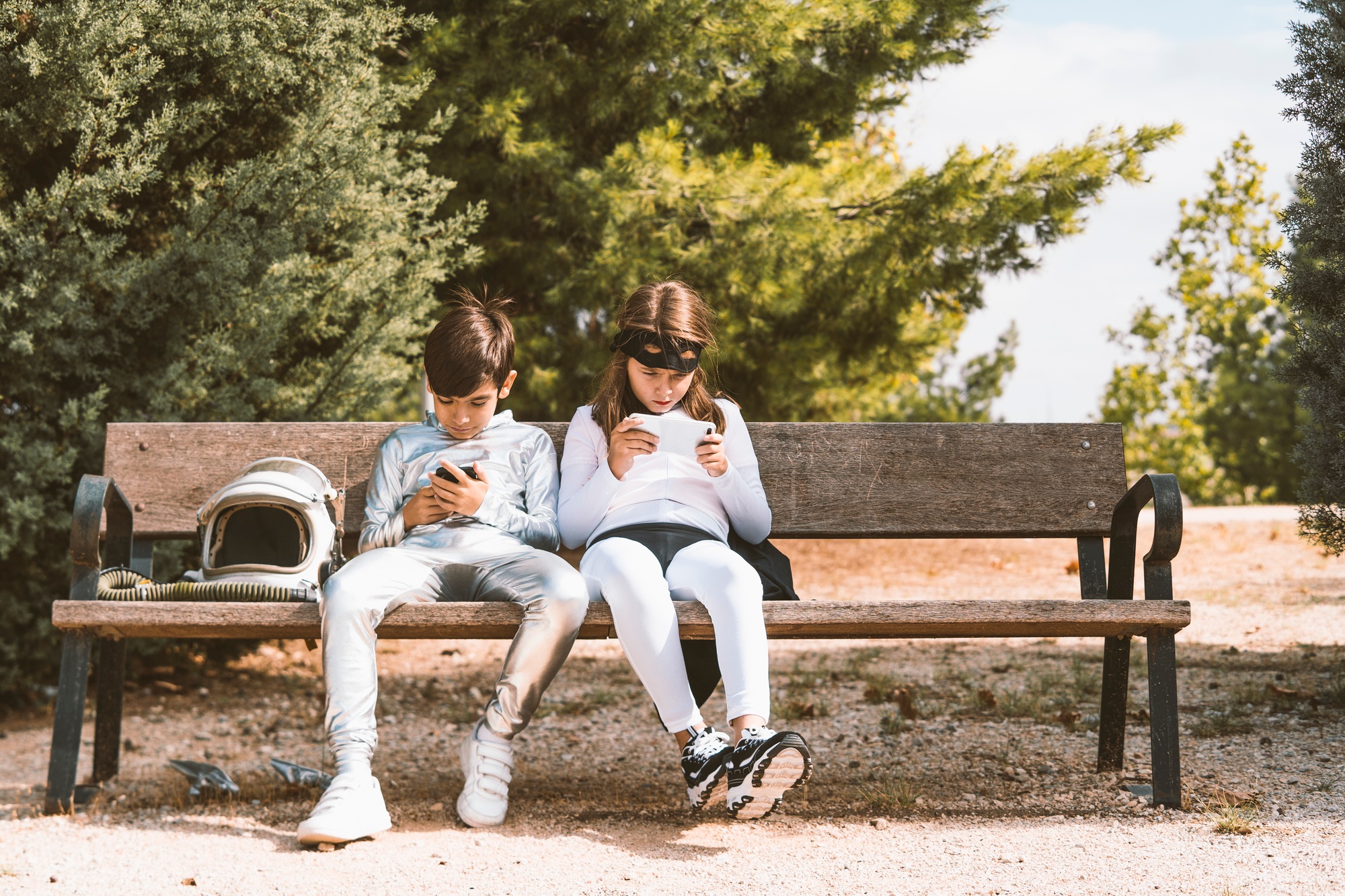 Two kids in astronaut and superhero costumes using mobile phone on park bench